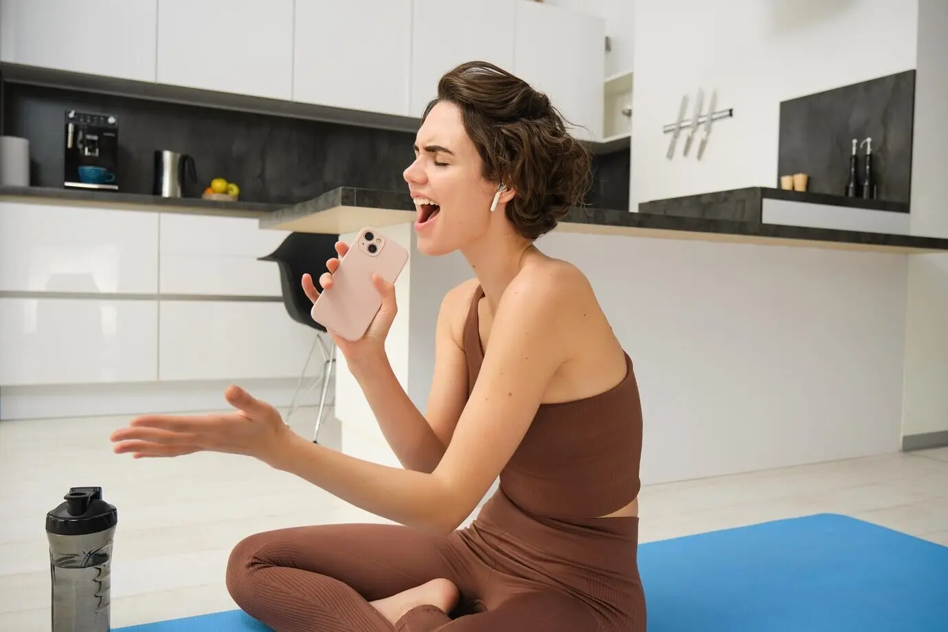 Unbeschwertes Fitness-Girl, Workout-Trainerin, sitzt zu Hause mit Wasserflasche und Yogamatte und trägt einen Trainingsanzug.